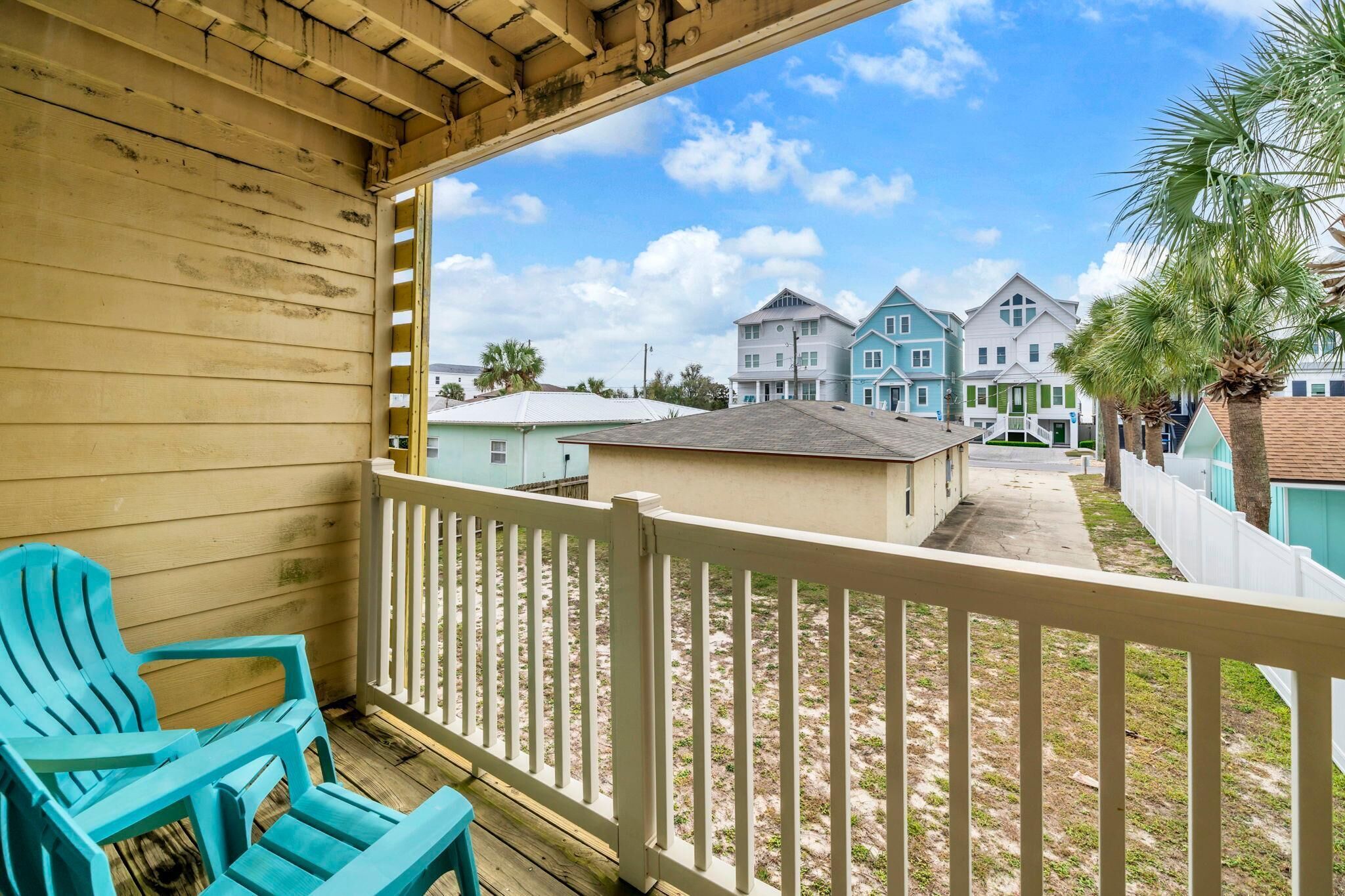 A wooden balcony with a white railing overlooking a residential area with colorful houses and a palm tree.
