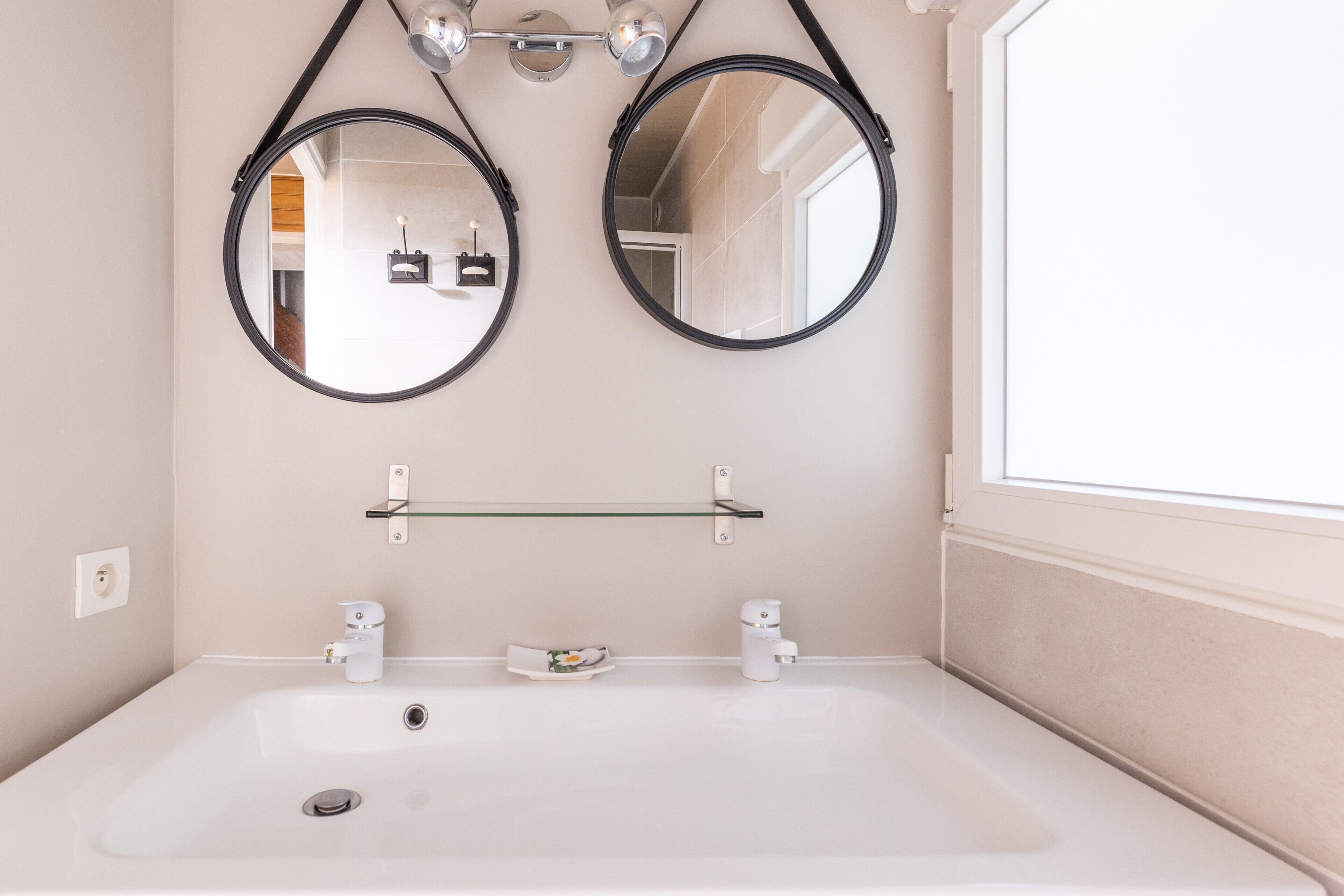 A bathroom with two round mirrors, a white sink, a glass shelf, and a window.