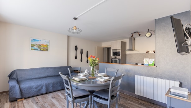 A dining area with a round table, chairs, a vase with flowers, a sofa, a kitchen in the background, and a wall-mounted clock. - Gîte à l'aube – 3 bedrooms (Saint-Aignan)