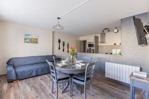 Ein Essbereich mit einem runden Tisch, Stühlen, einer Vase mit Blumen, einer Couch und einer Küche im Hintergrund sowie einer an der Wand befestigten Uhr.