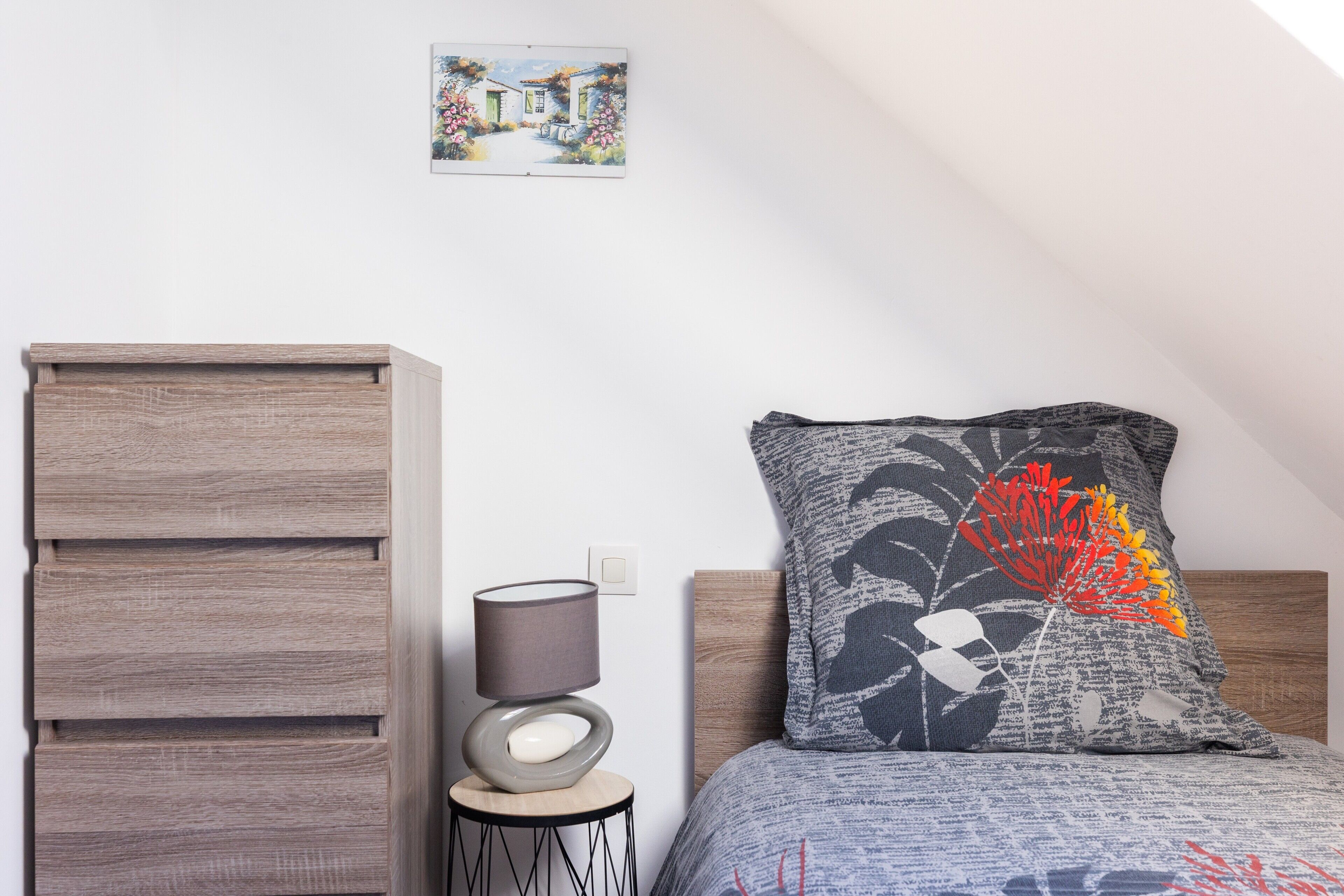 A bedroom with a wooden dresser, a bedside table with a lamp, a decorative pillow, and a framed picture on the wall.