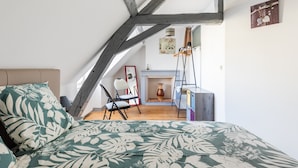 A bedroom with a sloped ceiling, a bed with a floral bedspread, a chair, a dresser, and a fireplace. - Gîte à l'aube – 3 bedrooms (Saint-Aignan)