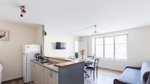 A compact kitchen with a refrigerator, coffee maker, and dining table with chairs. A flat-screen TV is mounted on the wall, and a large window provides natural light. - Gîte à l'aube – 3 bedrooms (Saint-Aignan)