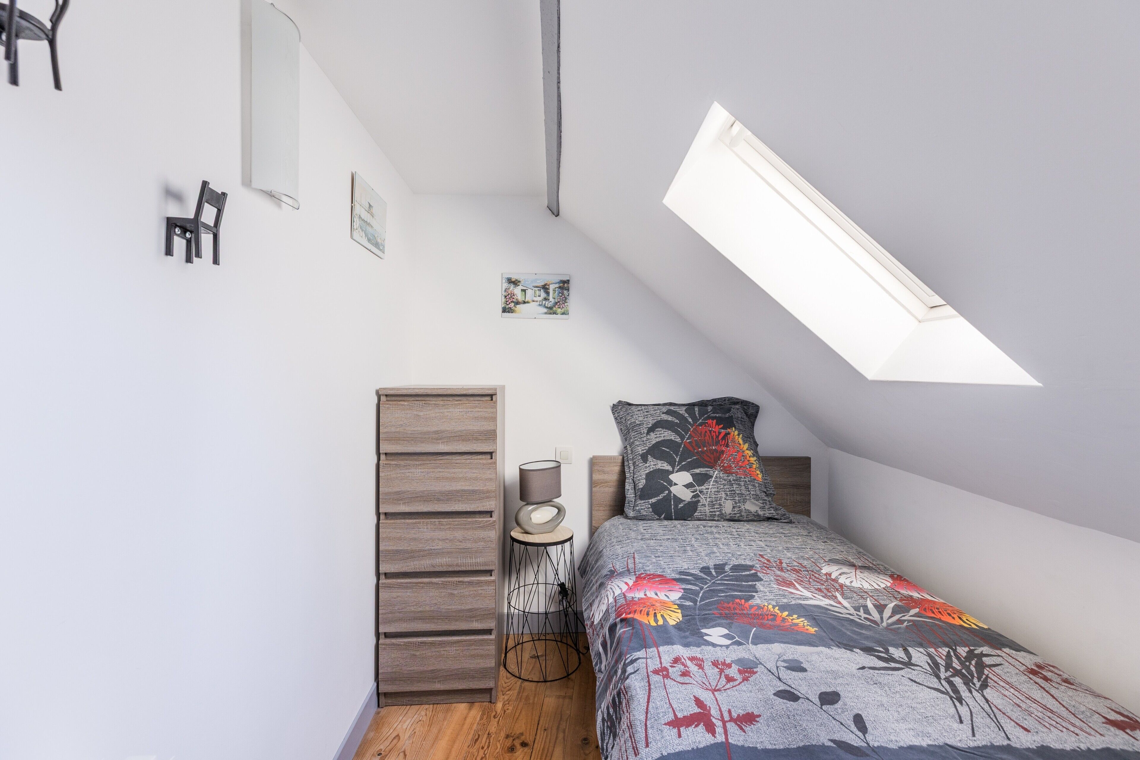 Une chambre avec un plafond en pente, un lit recouvert d’une couvre-lit à motifs floraux, une petite table de chevet, une armoire en bois et un puits de lumière.