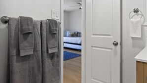 Une salle de bain avec deux serviettes suspendues à un porte-serviettes, une porte blanche et une chambre visible avec un lit et un tapis bleu.