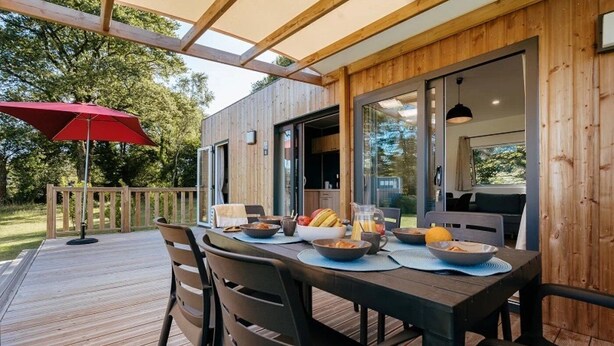 An outdoor dining area with a wooden deck, a table set for breakfast, and a red umbrella. - ZEcamping 4* espace bien-être avec jacuzzi et sauna cadre verdoyant (Caro)