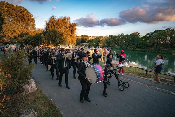 A group of musicians performing on a paved path by a body of water, with spectators in the background.