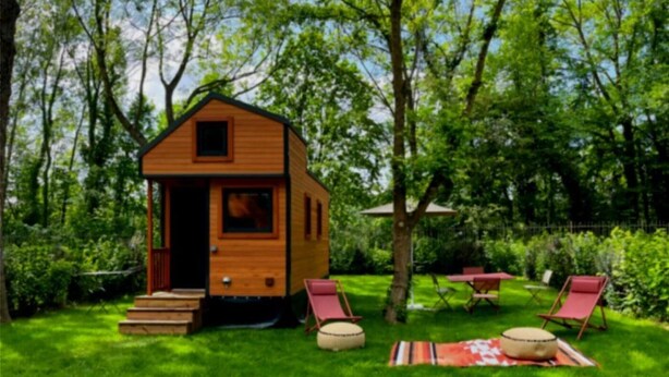 Eine kleine Holzhütte mit Veranda und Treppe, umgeben von üppiger Vegetation und Sitzgelegenheiten im Freien.