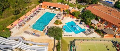 An aerial view of a resort with a large swimming pool, a water slide, a tennis court, and several lounge areas.