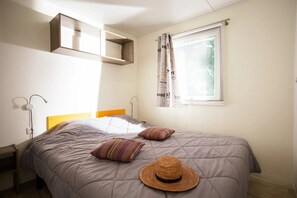 Ein Schlafzimmer mit einem Bett, zwei Kissen, einem Hut, einem Nachttisch, einem Wandregal und einem Fenster mit Vorhängen.
