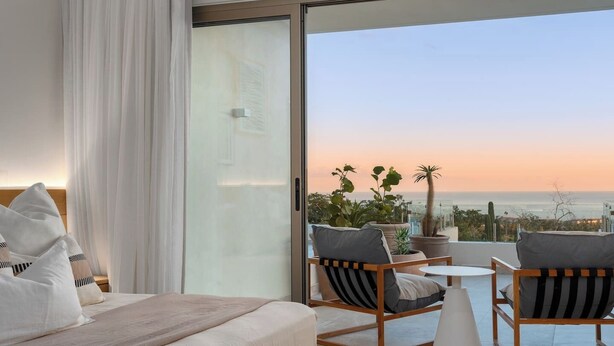 A hotel room with a bed, two chairs, a small table, and a view of the sunset over the ocean. - Cabo Costa - Private Infinity Pool and Ocean View (Cabo San Lucas)