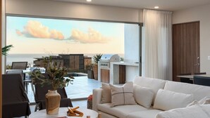 A modern living room with a large window overlooking a cityscape, a dining area with a table and chairs, and a kitchen area with a grill. - Cabo Costa - Private Infinity Pool and Ocean View (Cabo San Lucas)