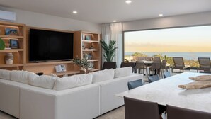 A modern living room with a large flat-screen TV, a white sofa, and a dining area with a table and chairs. There is a bookshelf with various items and a large window offering a view of the ocean. - Cabo Costa - Private Infinity Pool and Ocean View (Cabo San Lucas)