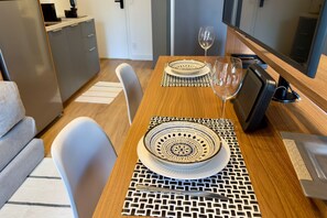 Een eetgedeelte met een houten tafel, witte stoelen, een decoratieve tafelmat, een schaal, een wijnglas en een televisie.