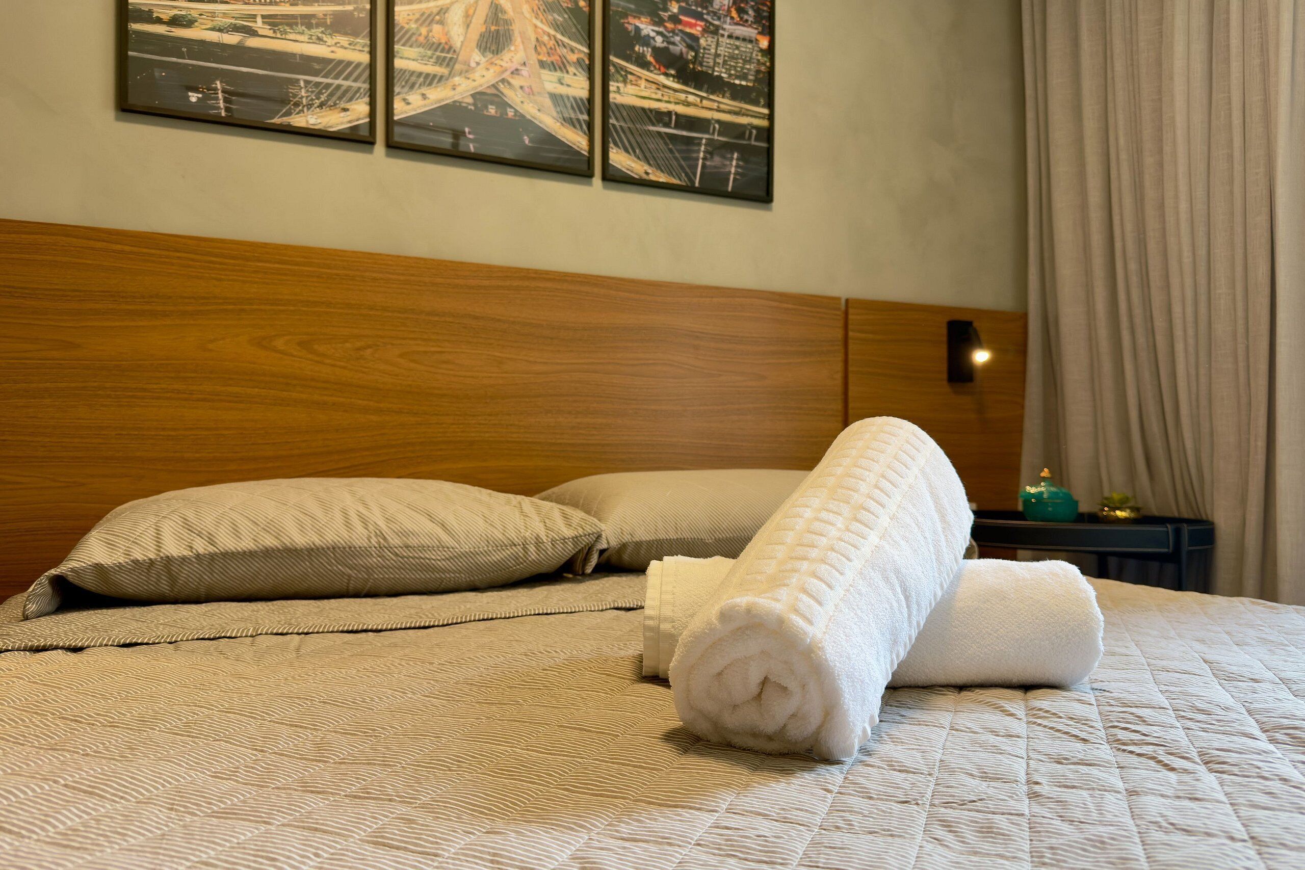 Una cama bien tendida con una toalla blanca enrollada, un cabecero de madera y una mesita de noche con una lámpara.