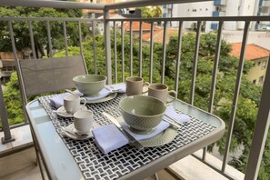 Un balcone con un tavolo apparecchiato per due persone, che include una ciotola verde, due tazze bianche e una tazza color beige.