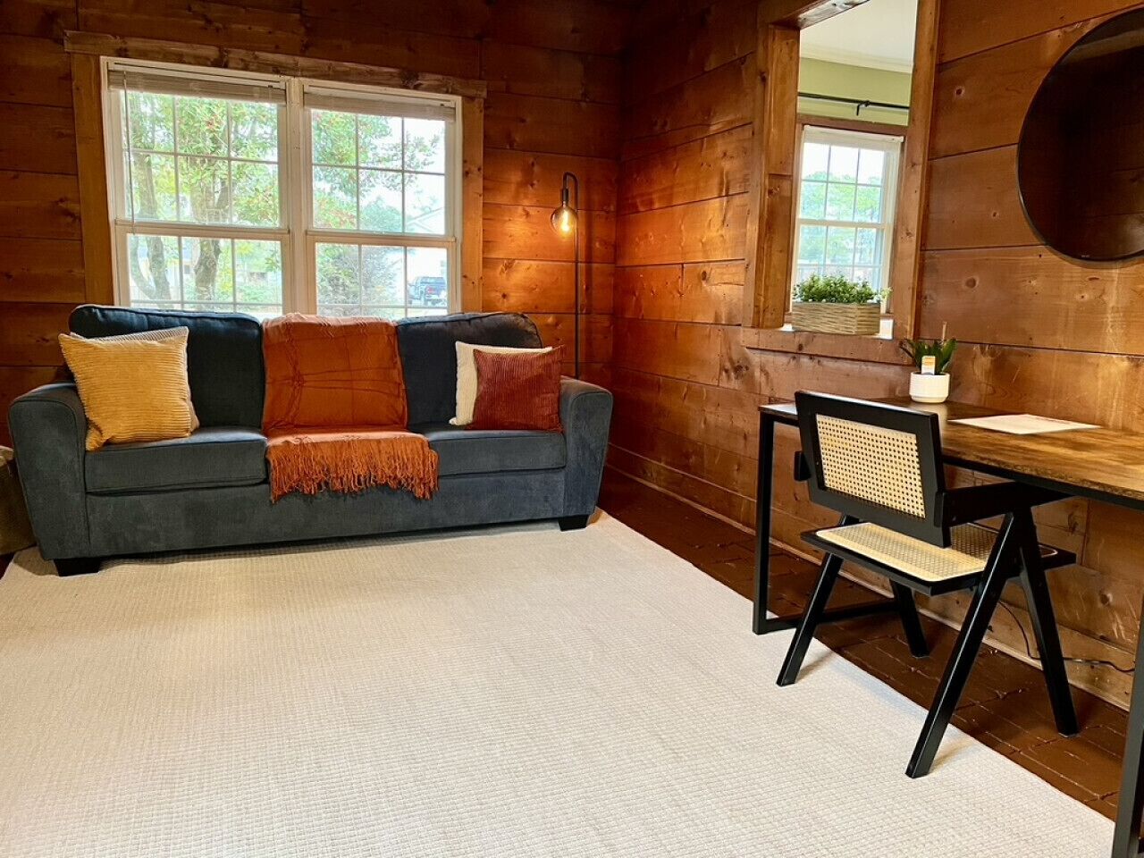 A cozy cabin interior with a sofa, a chair, a lamp, and a window with a view of trees.
