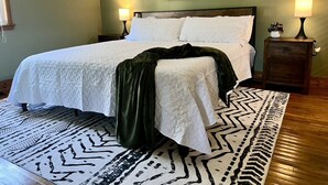 Una cama con ropa blanca, un plaid verde oscuro y una alfombra estampada.