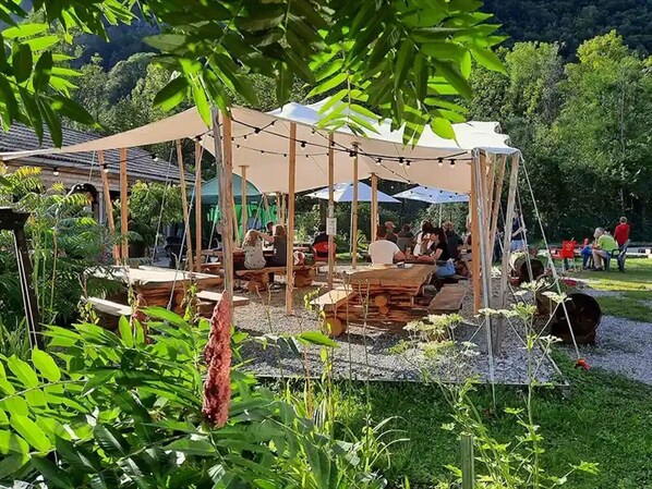 An outdoor seating area with tables and chairs under a canopy, surrounded by greenery.