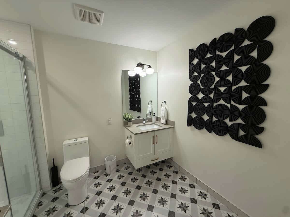 Une salle de bain avec des toilettes, un lavabo et un miroir. Il y a un espace douche avec des portes en verre. Le sol est recouvert de carreaux disposés selon un motif géométrique.