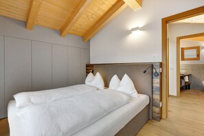 A bedroom with a wooden ceiling, a bed with white bedding, and a view into a bathroom. - Rostrini Family Home Alpine Retreat with Mountain View (Cavalese)