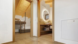 Quarto com cama beliche, teto de madeira, espelho redondo, pia e porta branca.