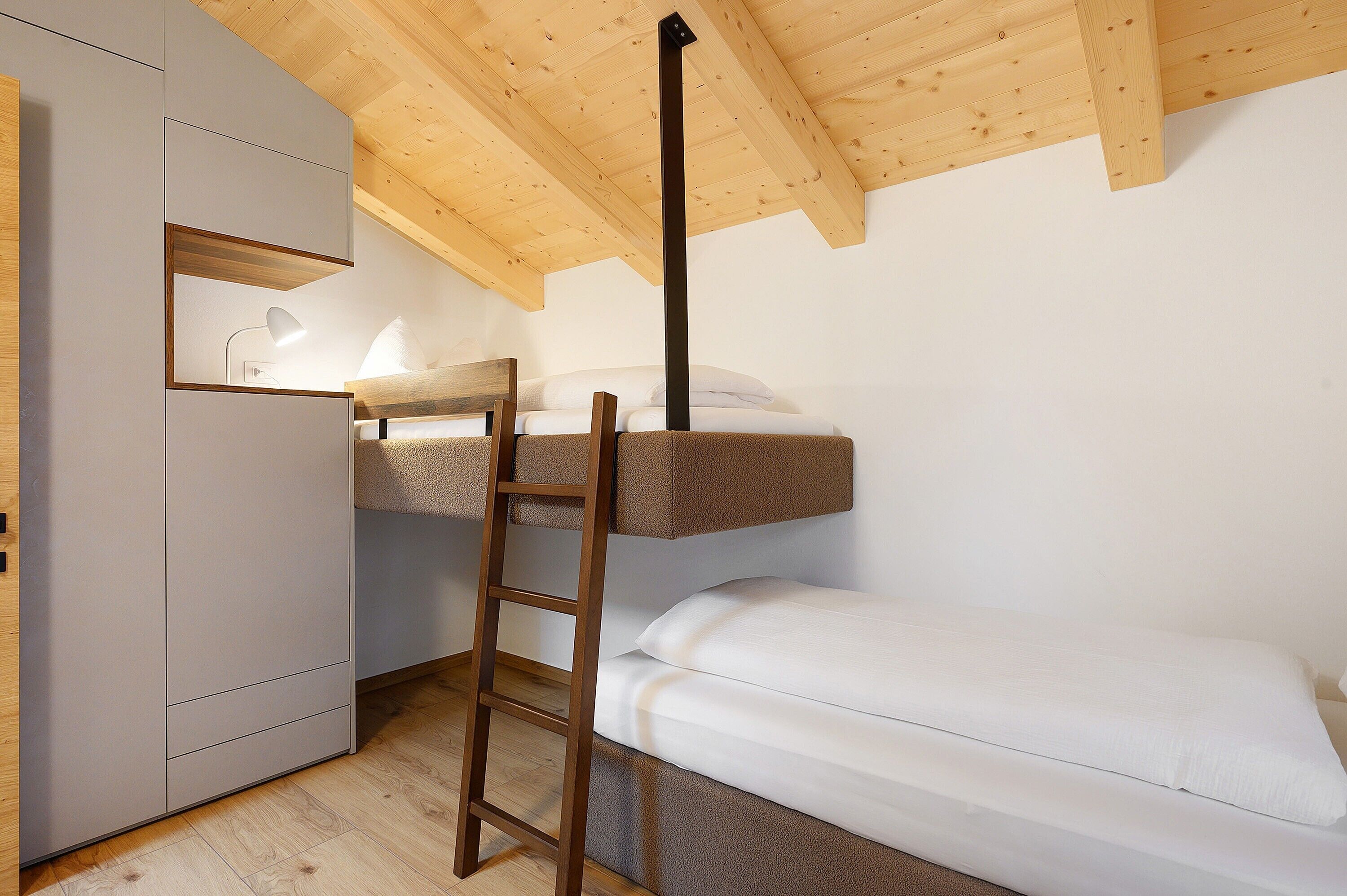 A bunk bed with a ladder, a desk with a lamp, and a wardrobe in a room with wooden ceiling beams.