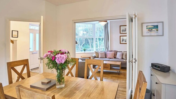 Een eethoek met een houten tafel, stoelen en een bloemenvaas. Achtergrond is een bank en een raam met gordijnen.