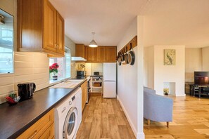 A compact kitchen with wooden cabinets, a washing machine, and a dining area with a grey sofa. - Host Stay Willow Way (Great Yarmouth)