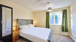 Ein Schlafzimmer mit einem großen Bett, einer hölzernen Kommode, einem Nachttisch, einem Fenster mit Vorhängen und einer Deckenleuchte.