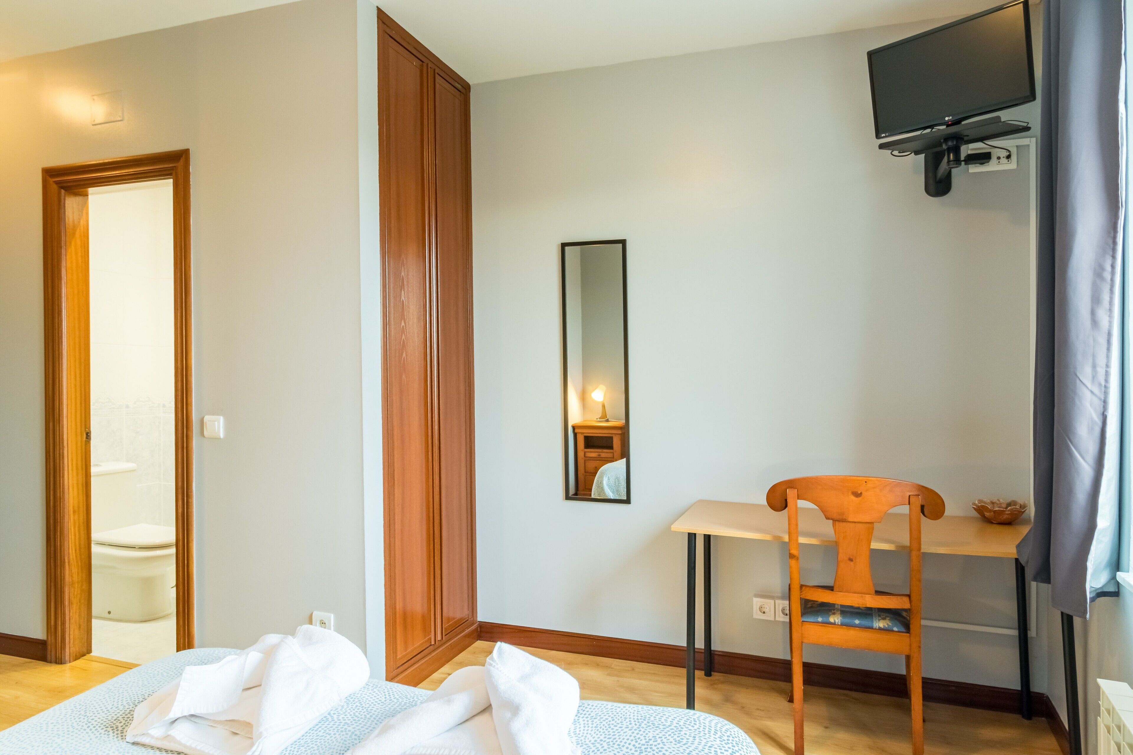 A bedroom with a bed, a wooden wardrobe, a desk with a chair, a television, and a mirror.