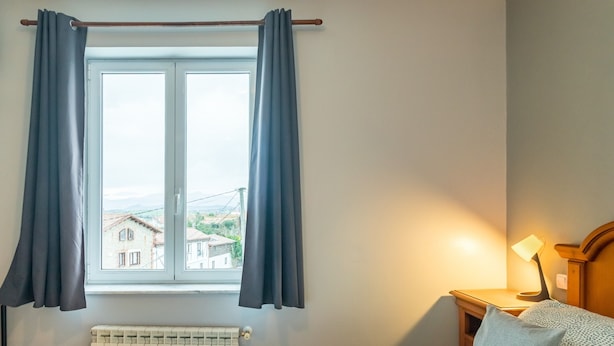 Een kamer met een bed, een raam met gordijnen, een radiator en een nachtkastje met een lamp.