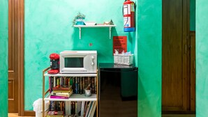 Een keuken met een magnetron, boekenplanken, een klein plankje met decoratieve objecten, een brandblusser en een koelkast.