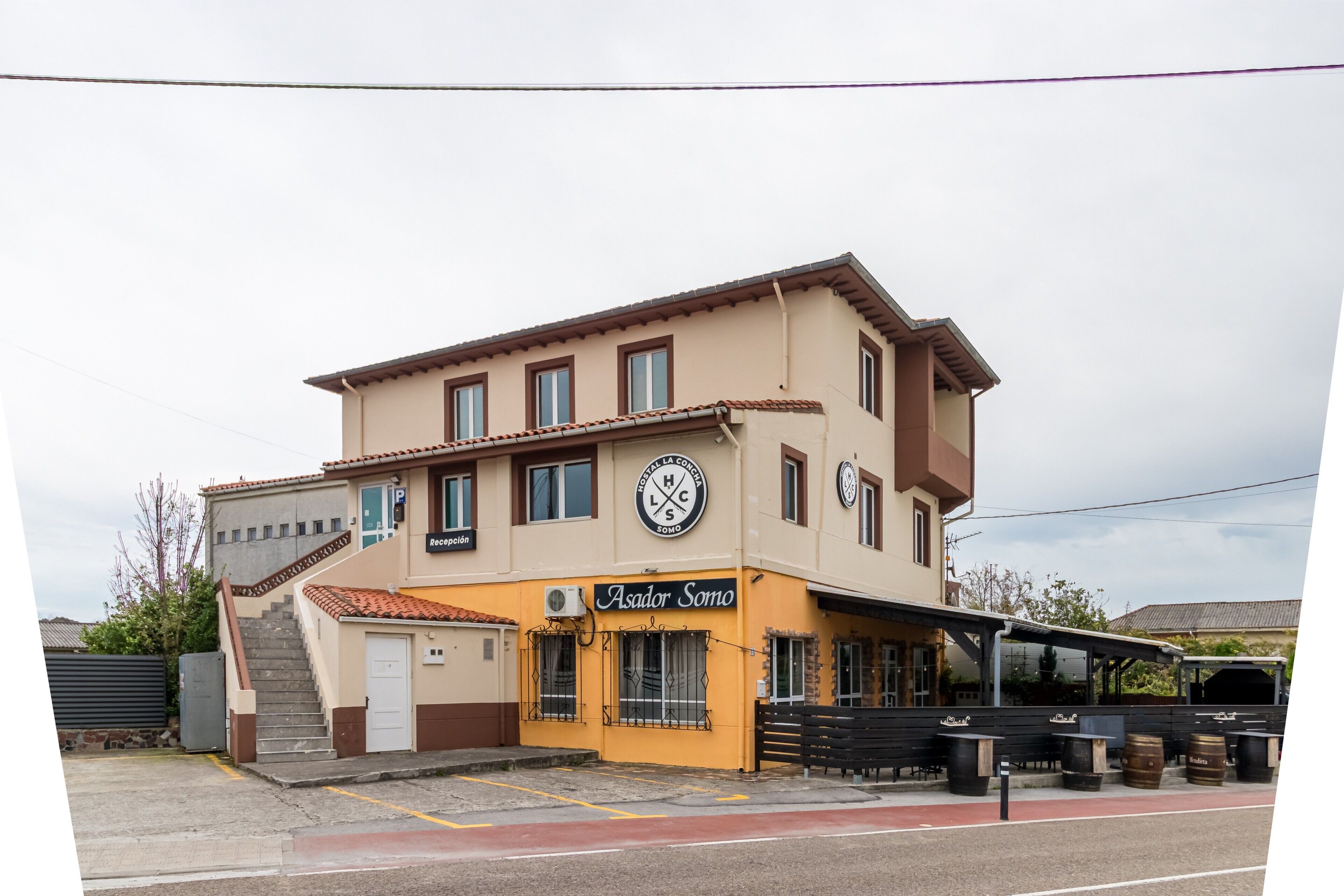 Un edificio a due piani con facciata gialla, un cartello con la scritta 'Asador Somo' e un'area di parcheggio con una fermata dell'autobus.
