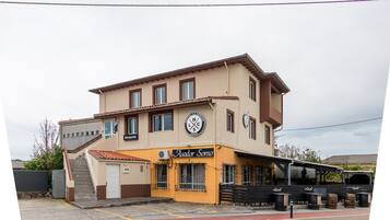Un edificio a due piani con facciata gialla, un cartello con la scritta 'Asador Somo' e un'area di parcheggio con una fermata dell'autobus.