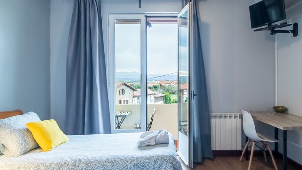 A bedroom with a bed, a desk, a chair, a TV, and a view of buildings through a window. - Habitación Doble 24 en Somo: Confort y Balcón Privado (Somo)