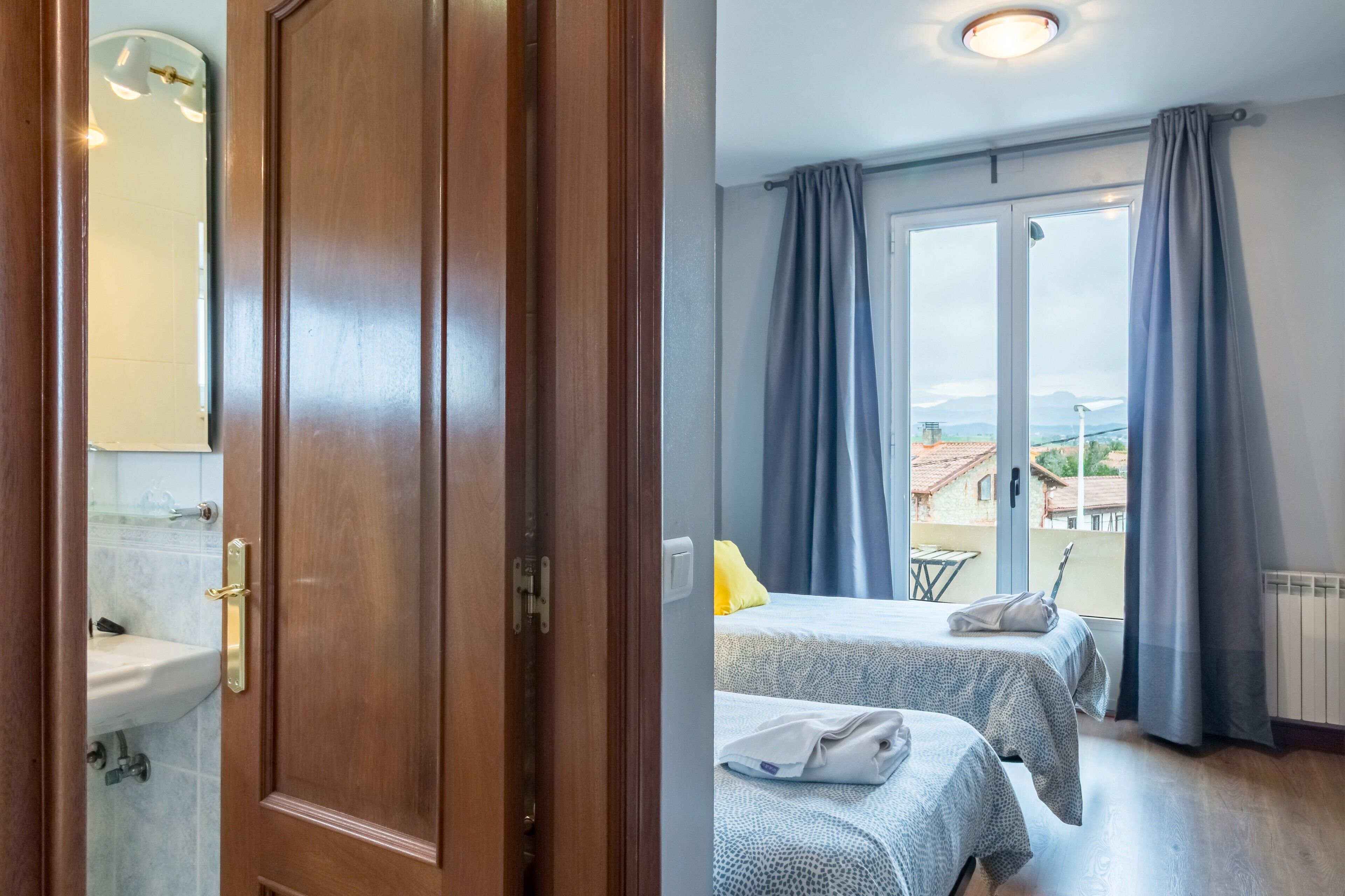 Une chambre d’hôtel avec une porte en bois, une salle de bain avec un lavabo et un miroir, deux lits avec une literie à motifs, et une vue dégagée sur l’extérieur grâce à de grandes fenêtres.