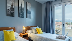A hotel room with two beds, a nightstand with a lamp, a window with a view of buildings, and three framed pictures on the wall. - Habitación Doble 24 en Somo: Confort y Balcón Privado (Somo)