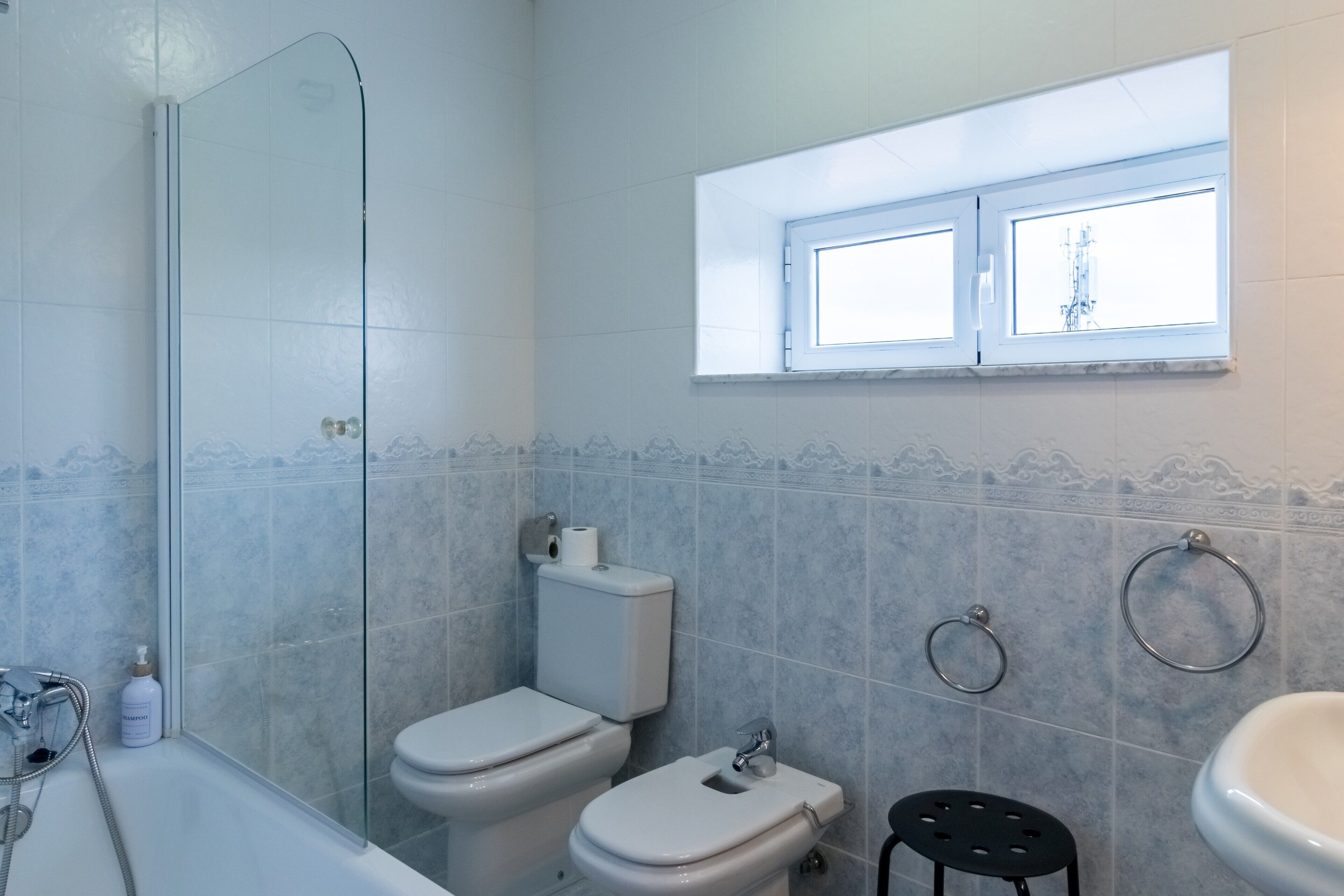 Ein Badezimmer mit Duschkabine aus Glas, zwei Toiletten, einem Waschbecken, einem Fenster und einem Handtuchhalter.