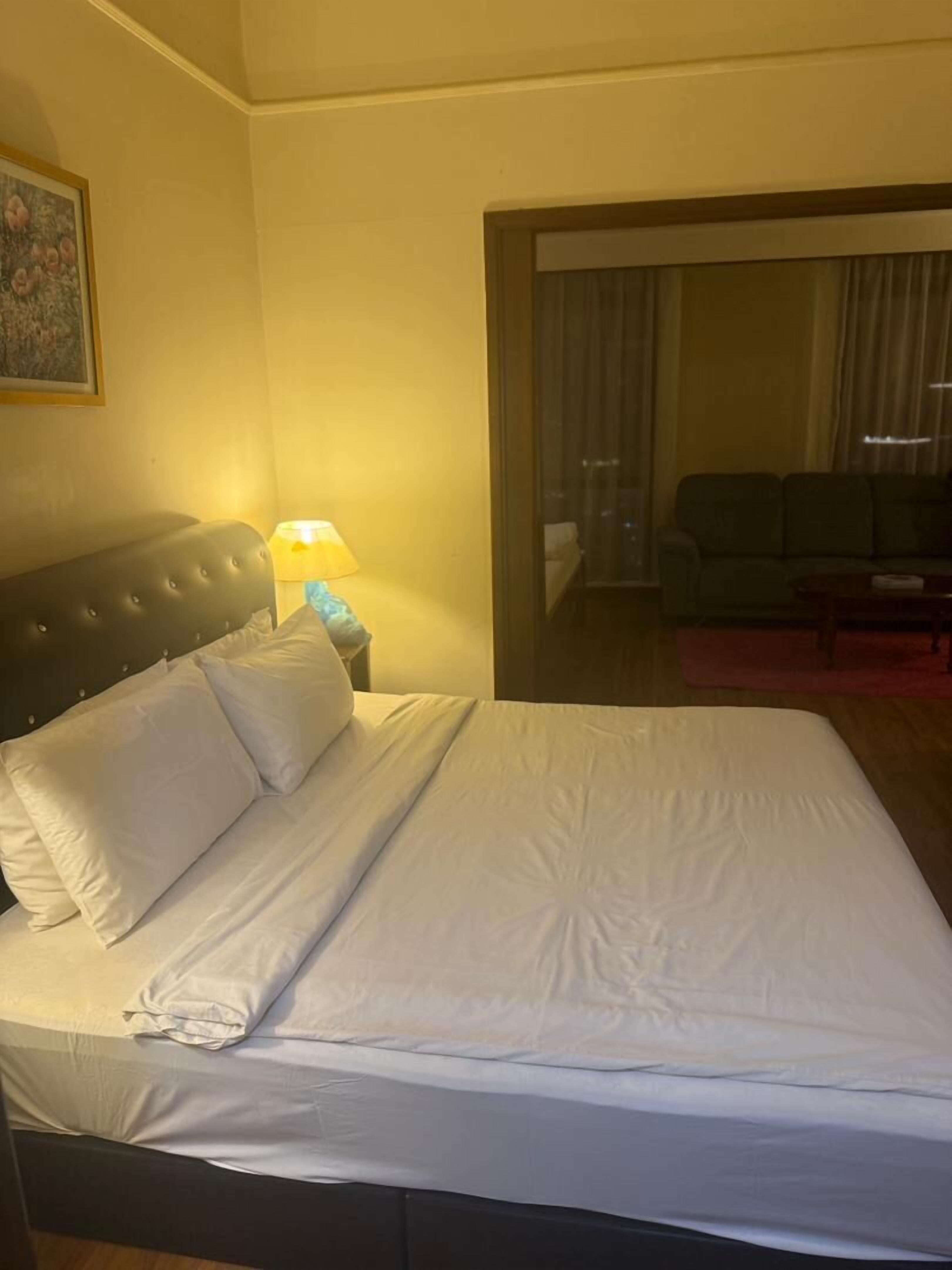 Habitación de hotel con cama, mesita de noche con lámpara, sofá y un amplio ventanal con cortinas.