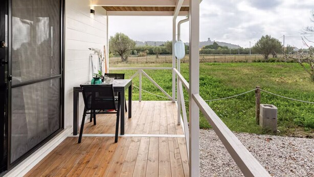 Un porche cubierto con una mesa y sillas de madera, vista a un campo verde y un edificio blanco con mosquitera.