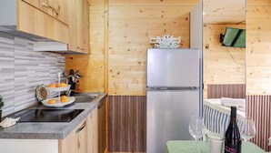 Una cocina compacta con armarios de madera, un refrigerador de acero inoxidable, un frutero de dos niveles y una pequeña zona de comedor con mesa y sillas.