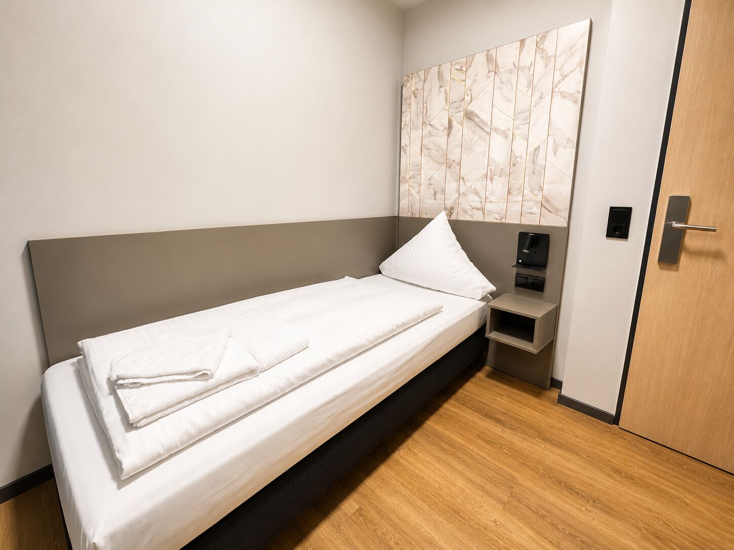 Ein Einzelbett mit weißen Laken, ein Nachttisch mit Ladestation und eine Holztür.