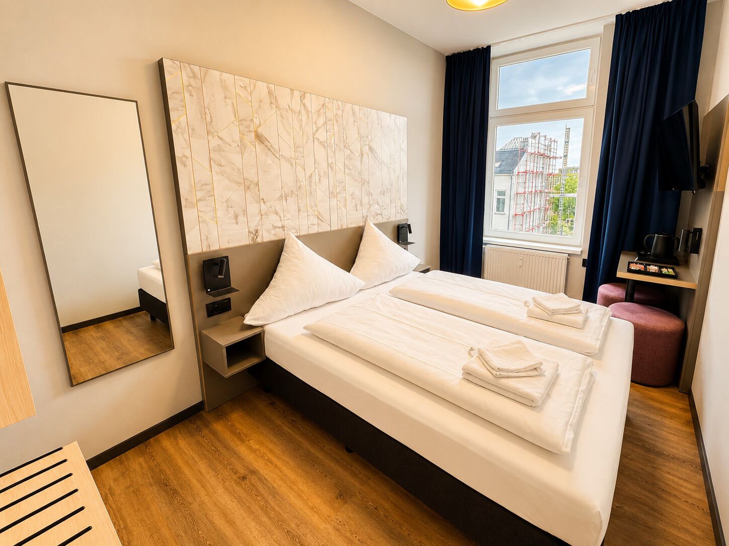 Habitación de hotel con una cama grande, una mesita de noche, un espejo, una ventana con vistas a edificios y una pequeña zona de estar.