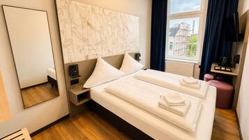 Ein Hotelzimmer mit einem großen Bett, einem Nachttisch, einem Spiegel, einem Fenster mit Blick auf Gebäude und einer kleinen Sitzecke.