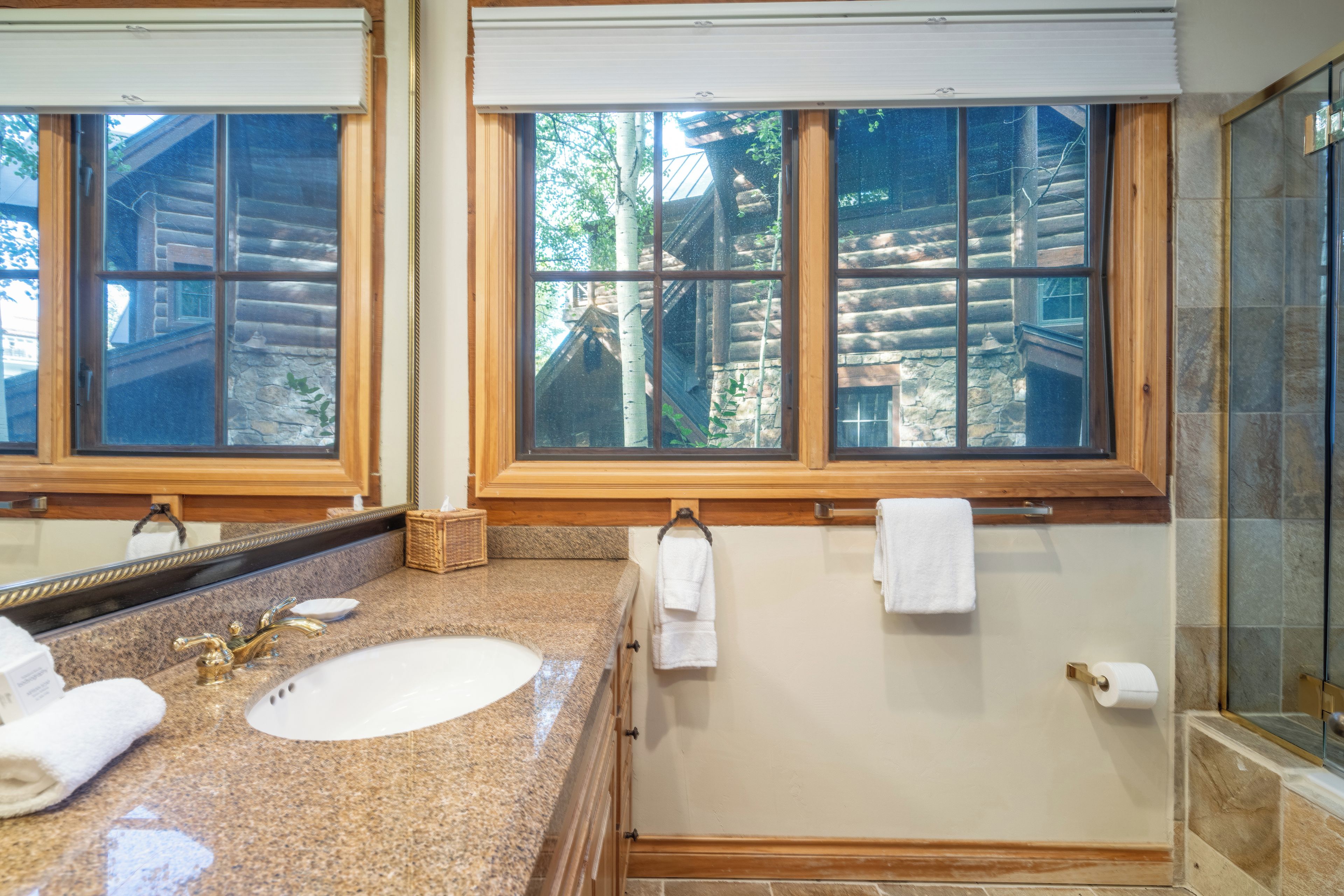 Un baño con encimera de granito, lavamanos, espejo y ventana con vista a una cabaña de madera.