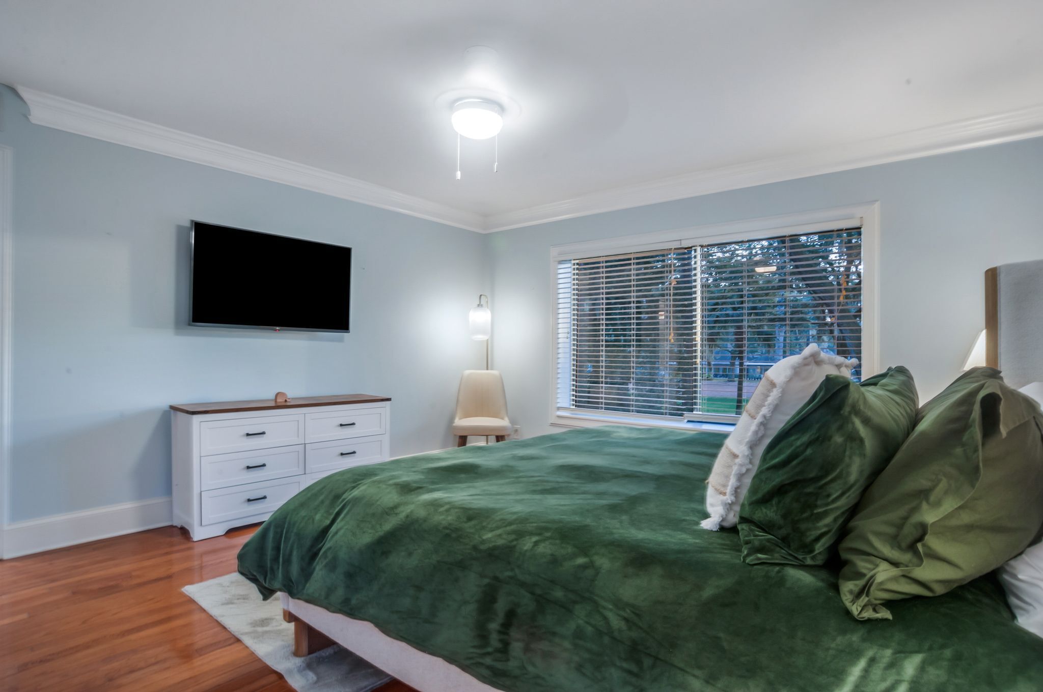 A bedroom with a bed, a dresser, a TV mounted on the wall, a window with blinds, and a nightstand with a lamp.