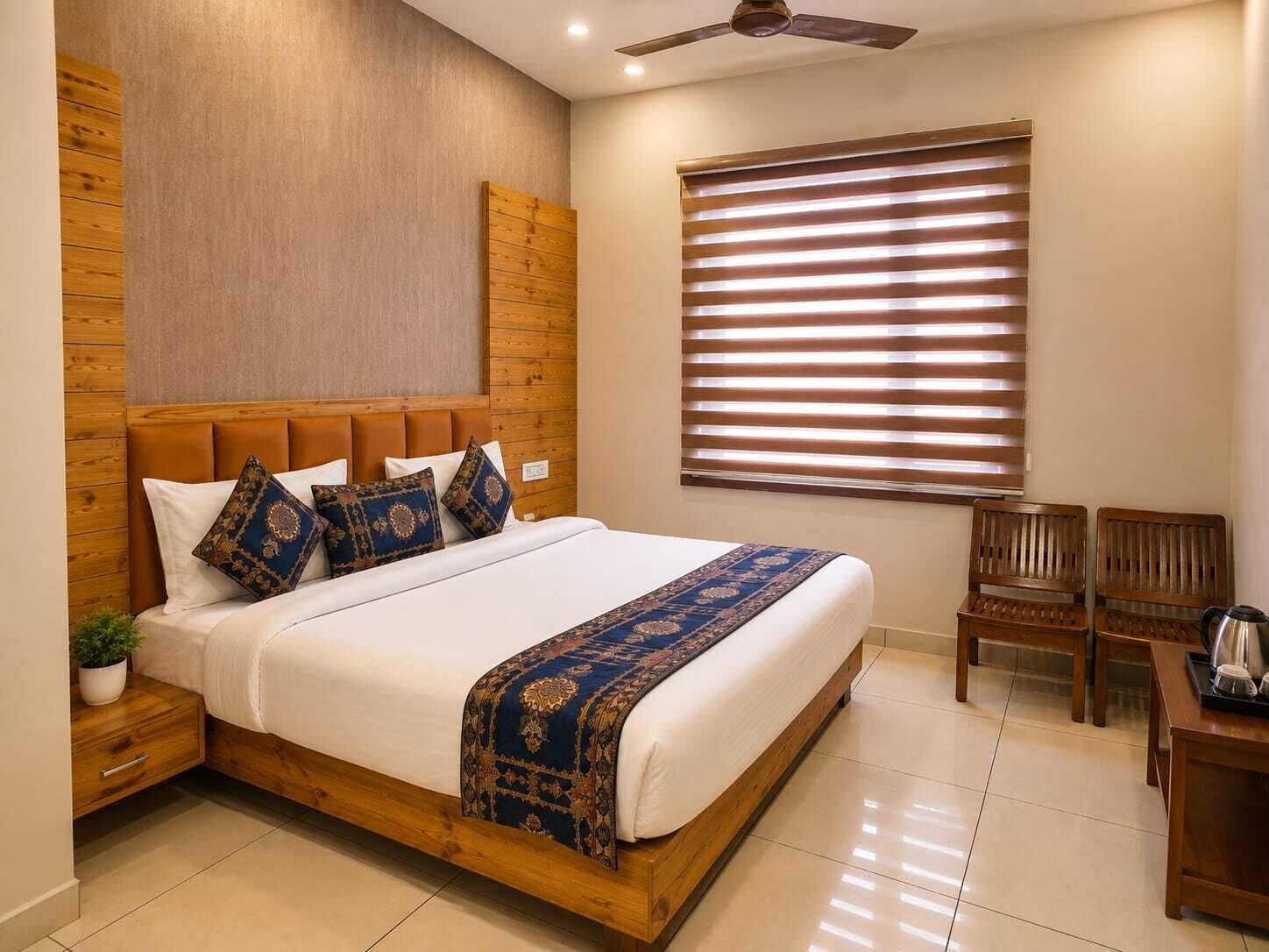 Habitación de hotel con cabecera de madera, cama grande con ropa de cama estampada, ventana con persianas, una mesita con hervidor y un ventilador de techo.