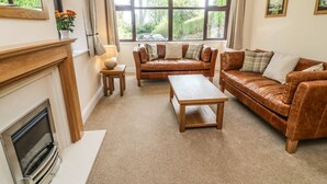 Una sala de estar acogedora con chimenea, sofás de cuero, una mesa de centro de madera y ventanas amplias que dan a vistas de vegetación.