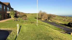 A grassy area with picnic tables and a flagpole, a building with a brick facade, and a road leading to a hilly landscape. - Cliffe Cottage (Saltburn-by-the-Sea)